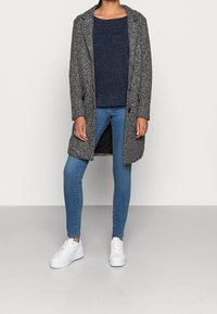 Abrigo texturizado gris, suéter de punto azul marino, vaqueros skinny azules y zapatillas blancas. El abrigo presenta un largo medio, detalles de botones y aberturas laterales.