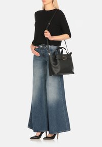 Borsa a mano in pelle nera con manici corti e una tracolla lunga e regolabile, con accenti in metallo dorato. Indossata con jeans a gamba larga blu chiaro.