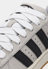 Világosszürke sportcipő fekete cikkcakk mintás oldalsávokkal, fehér, vastag, texturált fűzőkkel és egy bézs-krém színű talpélséggel, amely látható.