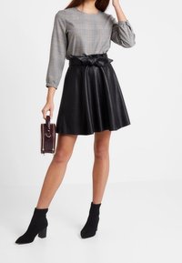 Haut à manches longues en coton gris à carreaux avec poignets froncés, jupe évasée en fausse cuir noire avec ceinture en nœud, et petit sac à main violet. Bottines noires.