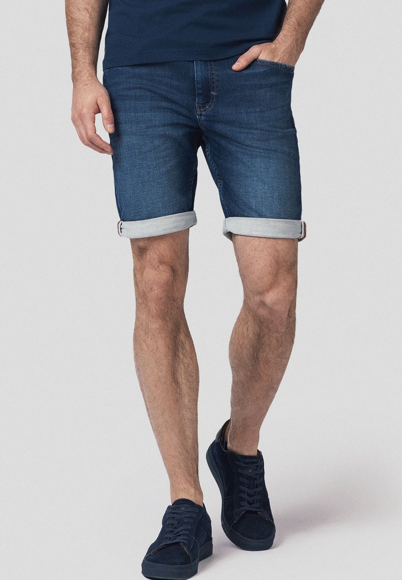 Jeansowe szorty w ciemnoniebieskim kolorze, z podwiniętymi mankietami, pięcioma kieszeniami i subtelnymi detalami w stichingu. W zestawieniu z granatowymi sneakersami.