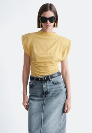 Kobieta nosząca ogromne czarne okulary przeciwsłoneczne, duże złote kolczyki koła, żółty top z poduszkami na ramionach oraz spódnicę z denimu z wysokim stanem i czarnym paskiem.