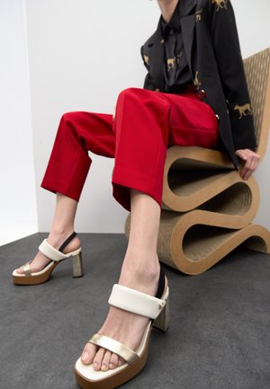 Zapatos de tacón alto con correas blancas y doradas, una base gruesa marrón, llevados con pantalones cortos rojos, sentada en una silla escultórica de cartón.