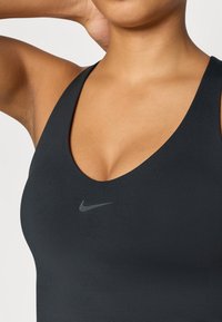 Černá sportovní podprsenka s hlubokým výstřihem do V, vyrobená z hladkého materiálu, s malým šedým logem Nike ve středu.