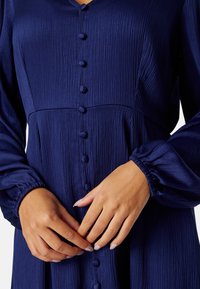 Tmavě modré, dlouhé šaty s texturovanou tkaninou, s knoflíkovým zapínáním vpředu a nařasenými manžetami. Model si upravuje knoflíky.