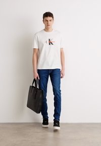 Hvit bomulls t-skjorte med svart og rød grafisk logo, blå denim jeans, svart lær tote-veske og svarte joggesko med hvite såler.