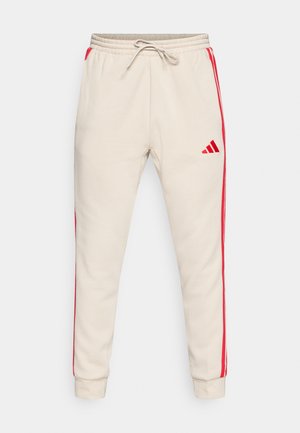 Calças de treino bege com riscas laterais vermelhas, cintura elástica com cordão e um pequeno logótipo vermelho na coxa esquerda. Textura suave.