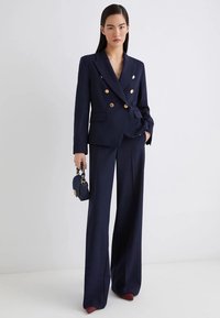 Blazer doppiopetto blu navy con bottoni dorati, abbinato a pantaloni blu a vita alta e una piccola borsa. Texture liscia e design sartoriale.