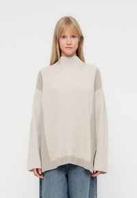 Pulover bej de mari dimensiuni cu guler înalt, textură ondulată și umeri căzuți. Purtați peste blugi largi de culoare albastru deschis.