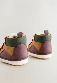 Ein Paar kleine, mehrfarbige Babyschuhe mit braunen, grünen und lila Elementen, roten Schnürsenkeln, weißen Sohlen und kuscheligem Innenfutter.