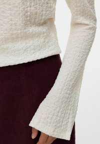 Cremig strukturierter Langarmtop mit ausgestellten Manschetten, kombiniert mit einem hoch taillierten, dunkelburgunderfarbenen Ripprock oder einer Hose.