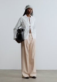 Fehér gombos ing nyakkendő részlettel, bézs színű bő szárú nadrág és fekete táska. A modell mintás fejkendőt visel. Simogató anyagtextúrák.