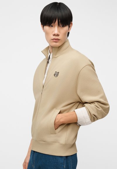 Νέος άνδρας φορώντας μπεζ ζακέτα με φερμουάρ και λογότυπο λύκου, με το χέρι στην τσέπη, κοιτάζει στο πλάι μπροστά σε απλό φόντο.
