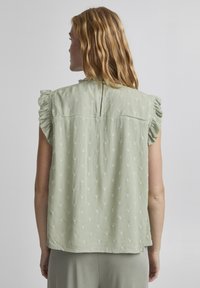 Blouse vert clair avec des manches à volants, en tissu texturé, et un motif à pois subtil. Détail de dos fendu. Design doux et décontracté.