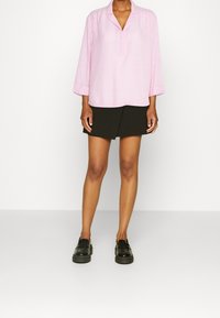 Hellrosa Bluse mit langen Ärmeln und Schlitzkragen, kombiniert mit einem schwarzen Wickelrock und schwarzen Loafers, die zusammen ein legeres Outfit ergeben.