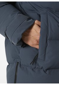 Detail na tmavě šedé péřové bundě, kde je vidět ruku, jak sahá do boční kapsy se zipem, která má strukturovanou, izolační látku.