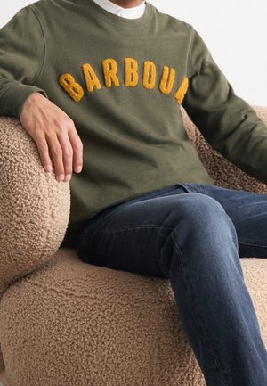 Osoba w oliwkowozielonej bluzie z dużym żółtym napisem „BARBOUR”, siedząca na beżowym, teksturowanym fotelu w niebieskich dżinsach.