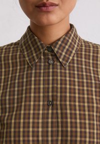 Chemise à carreaux brune et beige avec un col pointu, fermeture à boutons et tissu texturé. Comprend de petits boutons foncés et des coutures contrastées.