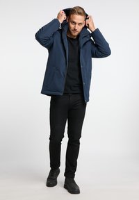 Tamsiai mėlynas izoliacinis paltas su užtrauktuku ir spaudėmis, reguliuojama kapišonu, dviem šoninėmis kišenėmis, uždėtas ant juodo marškinėlio ir juodų kelnių.