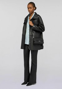 Giacca di pelle nera con grande colletto, chiusura a bottoni, indossata sopra una blusa azzurro chiaro. Pantaloni neri a zampa e scarpe nere a punta completano il look.