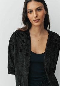 Black floral kimono-style jacket with short sleeves, featuring a textured pattern and a loose fit, worn over a dark tank top.