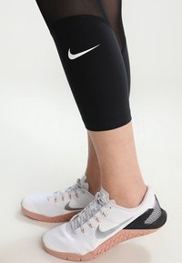 Black athletic capris with mesh details and a white Nike logo, paired with white and gray Nike fitness shoes featuring a pink sole.