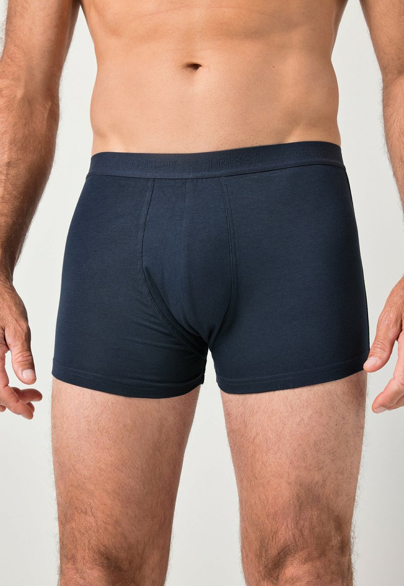 Männlicher Oberkörper und Oberschenkel, bekleidet mit schwarzen, eng anliegenden Boxer-Shorts mit elastischem Bund vor einfachem Hintergrund.