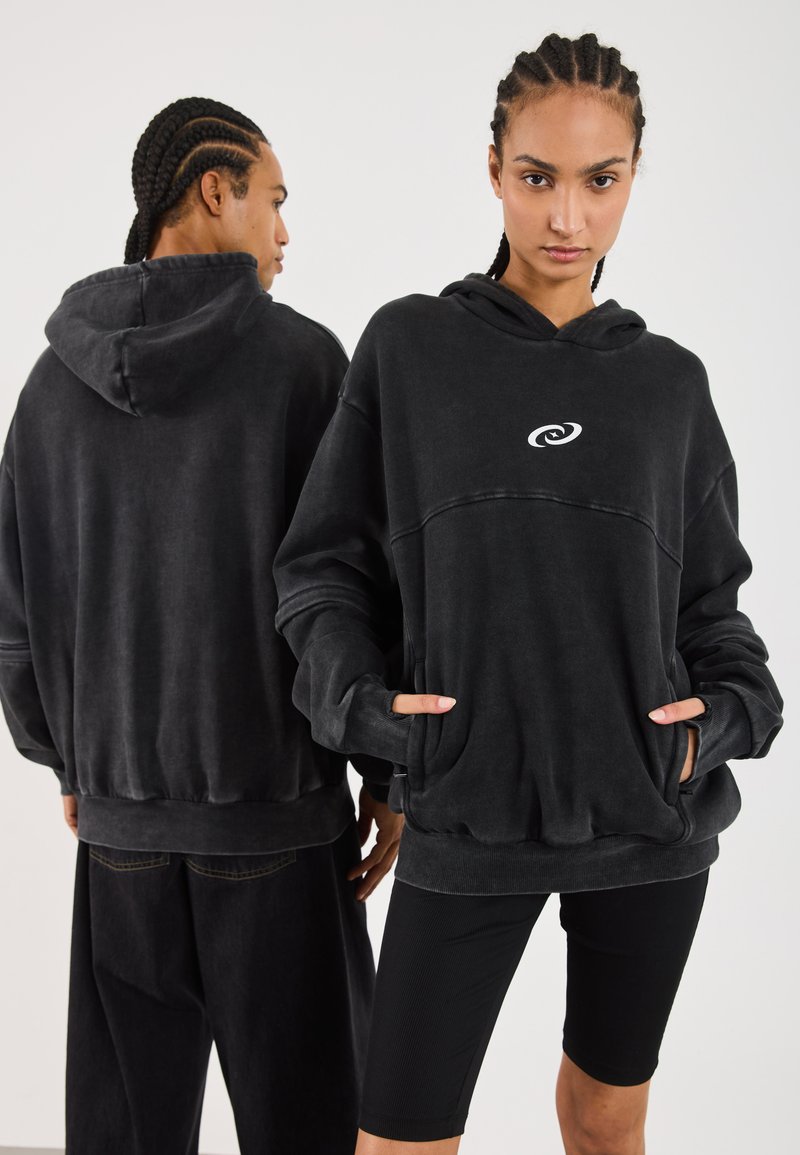 Felpa nera oversize realizzata in tessuto morbido, con una tasca frontale e un logo bianco. Il design ha una vestibilità rilassata e polsini a coste.