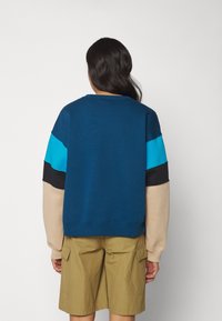 Felpa a blocchi di colore blu, nero e marrone chiaro con scollo rotondo e maniche lunghe, abbinata a pantaloncini beige con tasche cargo.
