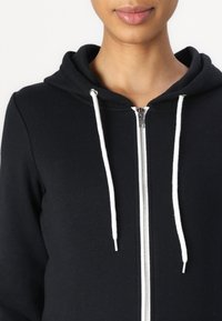 Sweat à capuche noir zippé avec un tissu texturé. Comprend une capuche à cordon avec des lacets blancs et une fermeture éclair argentée. Vue en gros plan.