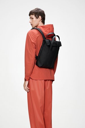 Črn nahrbtnik s strukturirano obliko, z dvema trakoma in zvitim zapiranjem na vrhu. Nošen čez oranžno vodoodporno jakno in hlače.