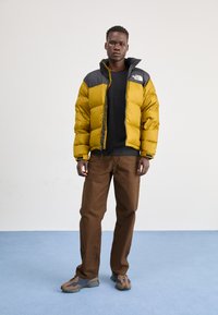Geel pufferjack met zwarte schouders, rits aan de voorkant en ribbelmanchetten; gecombineerd met een zwart shirt en bruine broek; sportschoenen met bruine en blauwe accenten.