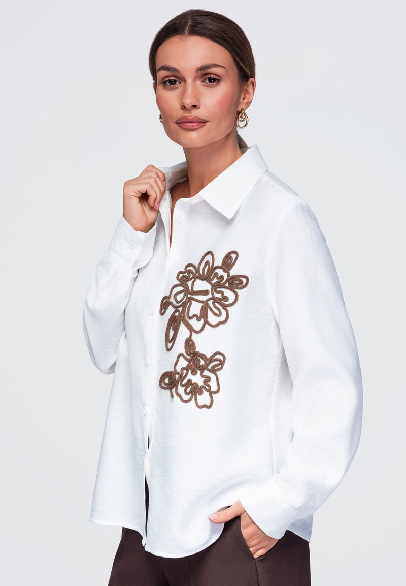 Femme aux cheveux bruns porte une chemise blanche à boutons avec une broderie florale marron, posant avec une main sur le col et l'autre dans la poche.