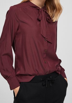 Hnědá košile na knoflíky s límečkem na zavazování, dlouhými rukávy a hladkou texturou; v kombinaci s černými kalhotami se stahovací šňůrkou.