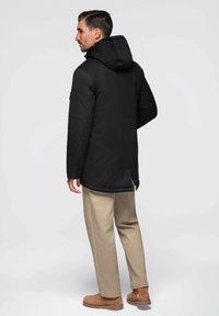 Chaqueta negra con capucha y textura acolchada, con parte trasera más larga, combinada con pantalones beige y zapatos marrones de apariencia ante.