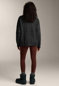 Person with curly hair wearing a black speckled sweater, burgundy leggings, gray socks, and black winter boots, standing facing away.