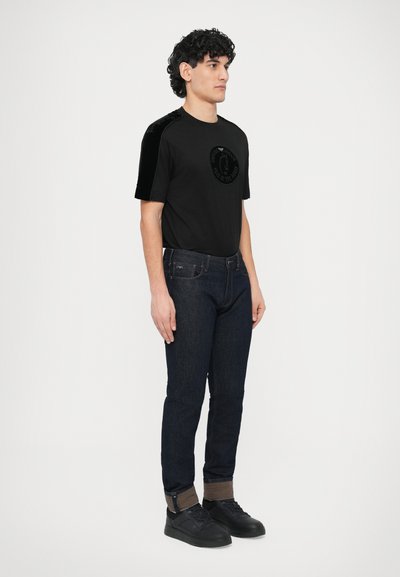 Fekete póló bársony vállrésszel és kör alakú logóval, sötét farmernadrággal és fekete tornacipővel párosítva. Egyszerű, testhezálló kialakítás.