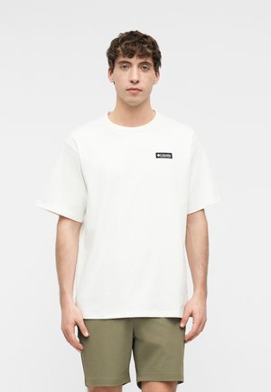 Junger Mann mit lockigem Haar, der ein weißes kurzärmeliges Hemd mit einem Logo und olivgrüne Shorts trägt, vor einem schlichten hellen Hintergrund steht.