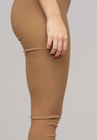 Testre simuló, barna leggings rugalmas anyagból. Sima textúrájú, oldalt varrott varrással rendelkezik. Kéz a lábon pihen.