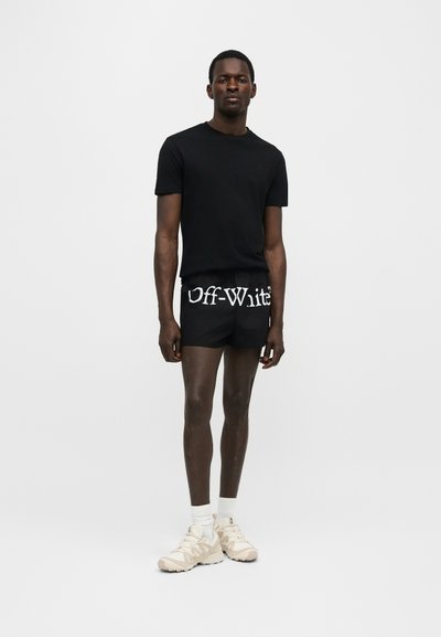 Homme portant un t-shirt noir à manches courtes et un short noir avec le logo blanc "Off-White", des chaussettes blanches et des baskets beiges, debout devant un fond uni.