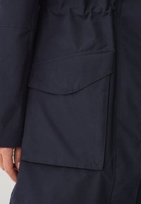 Donkerblauwe jas met een opvallende klepzak aan de linkerkant, gemaakt van een gladde stof met een matte afwerking. Eenvoudige stikdetails.