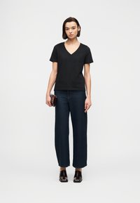 Γυναίκα με κοντά σκούρα μαλλιά που φοράει μαύρο T-shirt με V λαιμόκοψη, σκούρο τζιν με φαρδιά πόδια, σκούρα παπούτσια με τετράγωνη μύτη, κρατώντας ένα μικρό καφέ τσαντάκι.