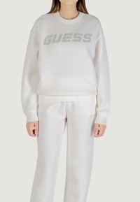 Biała bluza z bawełny oraz dopasowane spodnie z metalicznym logo "GUESS" na klatce piersiowej. Posiada ściągacze przy mankietach i w pasie.