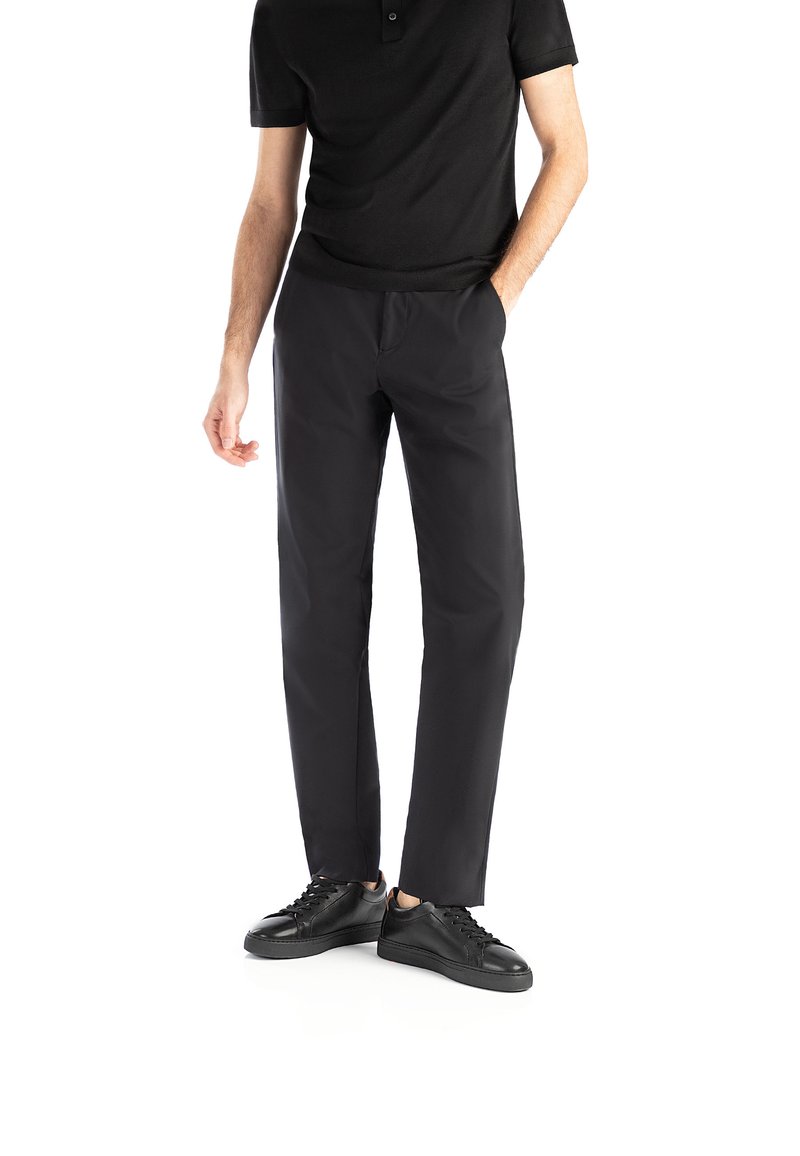 Uomo con indosso una polo nera, pantaloni neri e sneaker nere, in piedi con una mano in tasca su sfondo bianco.