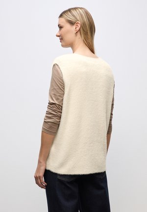 Top in maglia senza maniche color avorio con una finitura morbida e texturizzata. Ha un colletto rotondo ed è indossato sopra una camicia marrone a maniche lunghe.