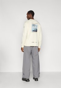 Μπλούζα μακρυμάνικη σε εκρού με γραφικό στο πίσω μέρος, γκρι φαρδιά παντελόνια και μαύρα παπούτσια με κορδόνια. Το κείμενο στην μπλούζα γράφει «Το μέλλον μας είναι η ΦΥΣΗ.»