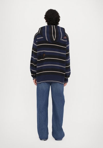 Sweatshirt à capuche rayé en bleu marine, noir et crème, présentant une coupe ample et un détail usé au dos. Associé à un jean bleu à jambes larges.