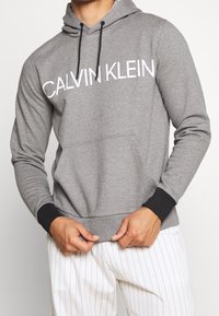 Sweat à capuche gris avec le logo "CALVIN KLEIN" en blanc, présentant une poche avant et des poignets côtelés noirs. Tissu doux et texturé.