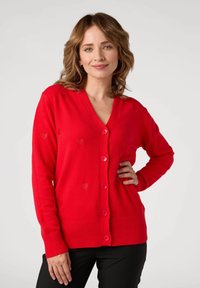 Cardigan rosso con scollo a V, decorato con motivi a cuore e bottoni rosa. Maniche a costine e orlo. Il materiale sembra morbido e lavorato a maglia.
