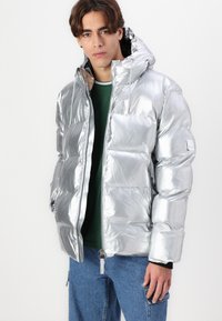 Giacca puffer argento con una finitura metallica lucida, dotata di cappuccio, chiusura con zip e tasche laterali. Indossata sopra un top verde e jeans blu.